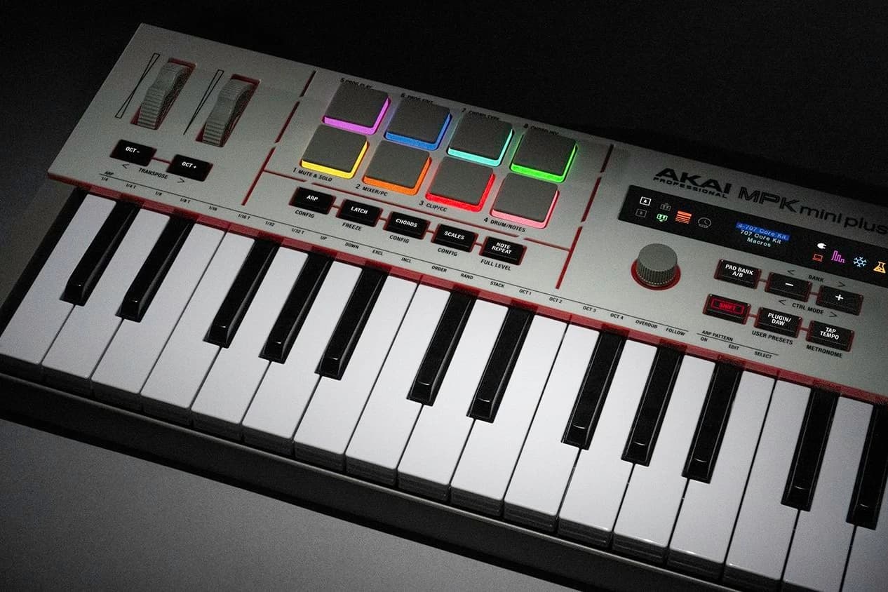 Jasnoszary kontroler Akai MPK mini plus z czerwonymi akcentami, widziany pod kątem w półmroku: osiem padów świeci różnymi kolorami, niżej białe i czarne klawisze, po prawej pokrętło i ekran z kolorowymi ikonami.