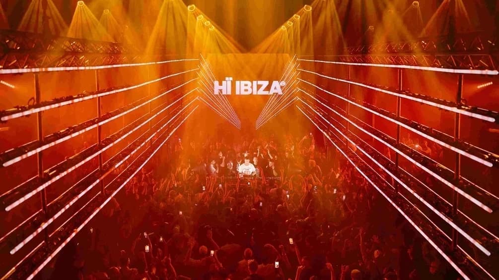 Wnętrze klubu Hï Ibiza w trakcie imprezy: tłum wokół centralnego boothu DJ-skiego, nad parkietem świecące logo HÏ IBIZA na ekranie LED, sala w fioletowym świetle