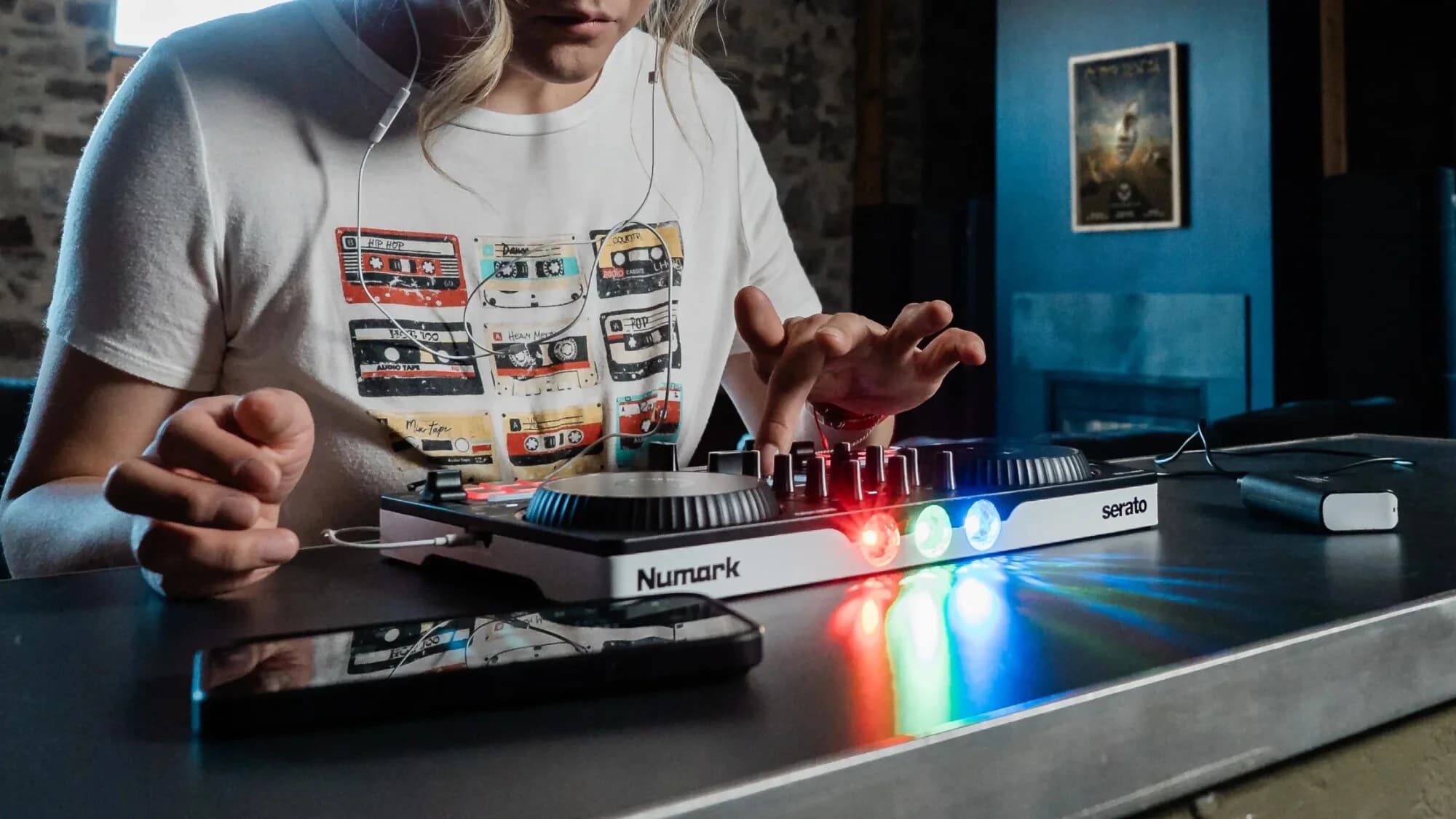 Osoba w białej koszulce z nadrukiem kaset magnetofonowych gra na kontrolerze Numark Party Mix III, z którego świecą czerwona, zielona i niebieska lampka; obok na stole leży smartfon
