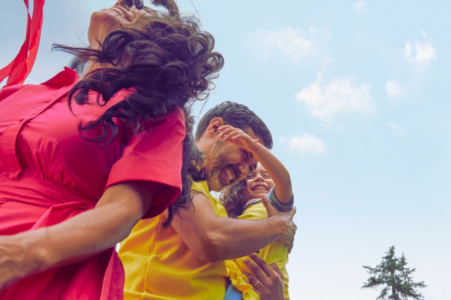 Zdjęcie Spotify promujące plan Premium Family: roześmiana dorosła osoba w żółtej koszuli trzyma na rękach dziecko, obok druga osoba w czerwonej sukience z rozwianymi włosami, w tle błękitne niebo