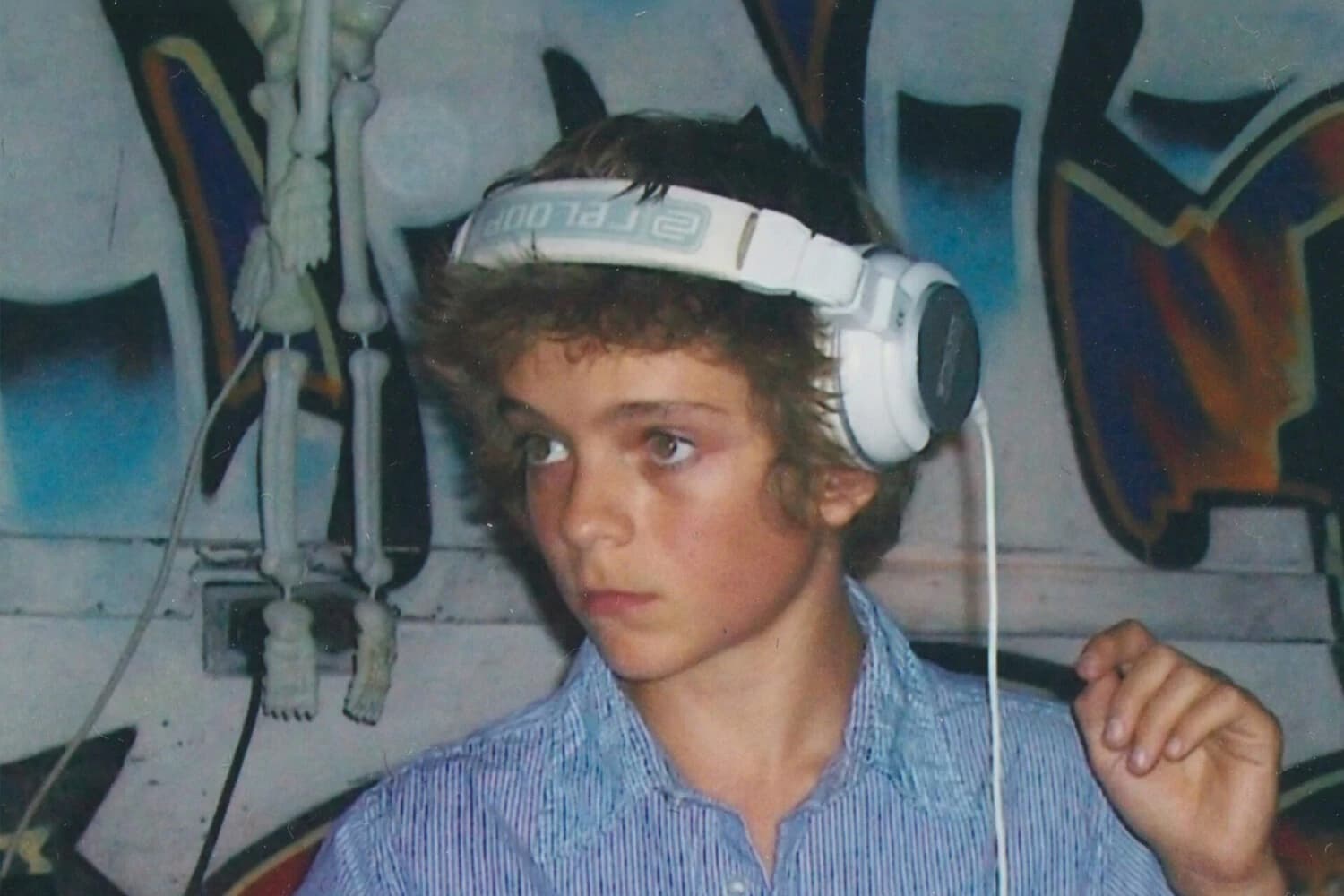 Zdjęcie z okładki albumu „MARTY”: kilkunastoletnia osoba z kręconymi włosami, w dużych białych słuchawkach i koszuli w niebiesko-białe prążki, na tle ściany z kolorowym graffiti i wiszącej obok dekoracji w kształcie szkieletu
