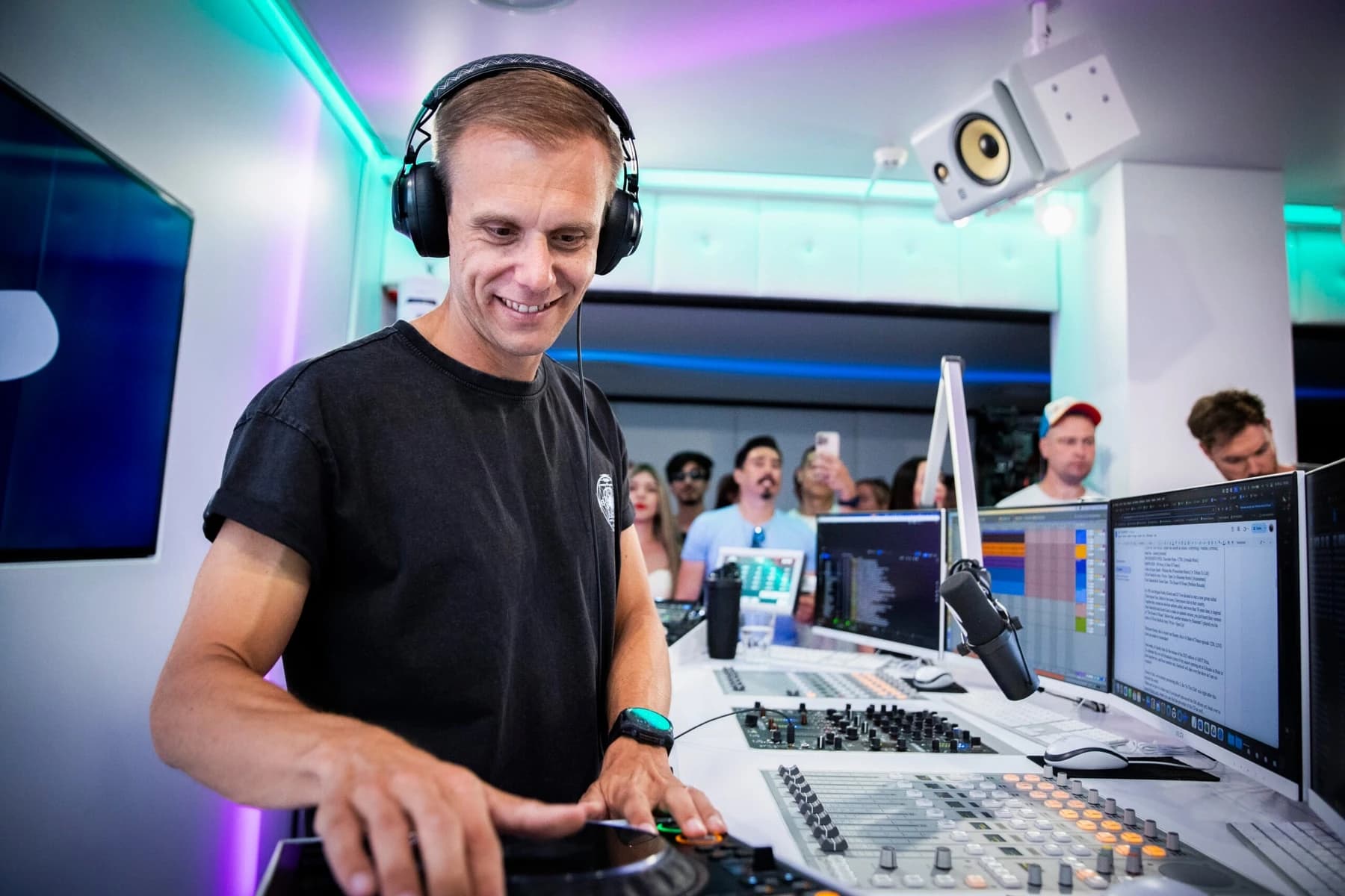 Uśmiechnięty, krótko ostrzyżony mężczyzna w czarnej koszulce i słuchawkach pochyla się nad kontrolerem DJ w jasnym studiu radiowym z konsoletą i monitorami, za nim grupa osób, w turkusowym i fioletowym świetle.