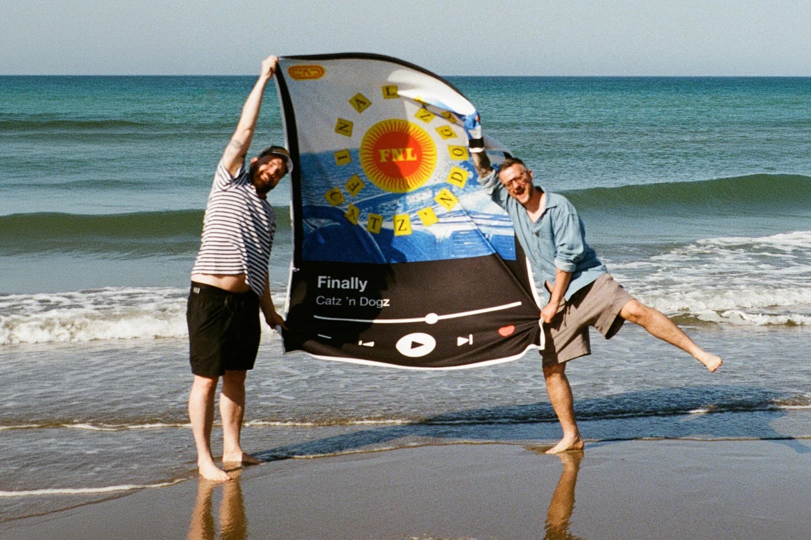 Catz ’n Dogz na plaży trzymają między sobą rozpięty ręcznik z okładką albumu „Finally”, w tle morze