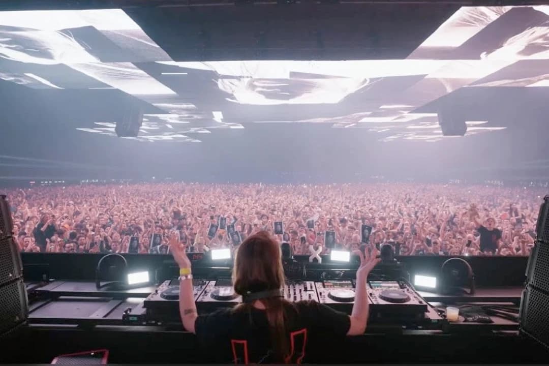 Charlotte de Witte ujęta od tyłu, z uniesionymi rękami za konsoletą DJ-ską, przed tłumem tysięcy osób w hali z podświetlonym sufitem; część publiczności trzyma w górze kartki z portretami