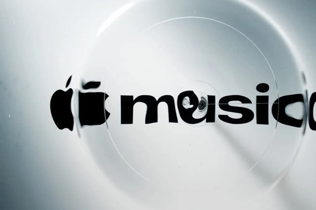 Czarne logo Apple Music z jabłkiem i napisem „music” widziane przez przezroczysty krążek, który zniekształca litery, na srebrzystobiałym tle w chłodnym świetle.