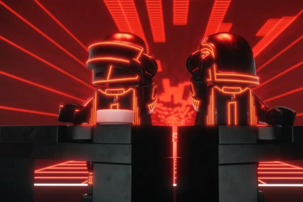 Dwie figurki LEGO Daft Punk w czarnych hełmach i strojach obrysowanych pomarańczowym neonem stoją za klockowymi pultami na tle czerwonej sceny z promieniującymi liniami światła.
